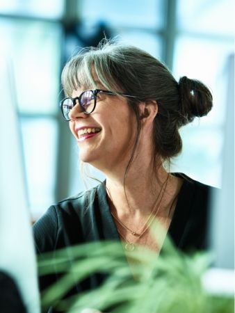 A woman over 50 wearing glasses, looking at a computer screen and smiling