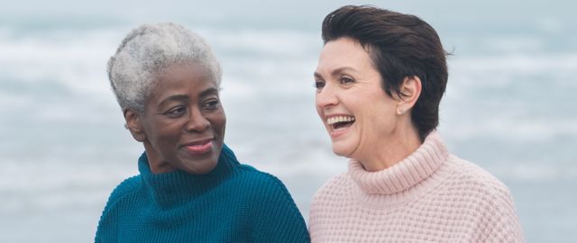 Two women over 50 wearing jumpers, walking and talking on a beach