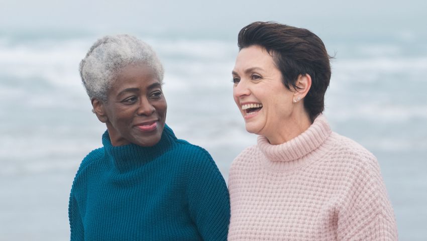 Two women over 50 wearing jumpers, walking and talking on a beach