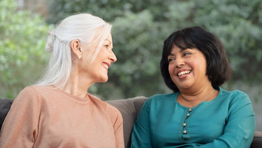 Two woman over 50 sitting on a sofa smiling at each other
