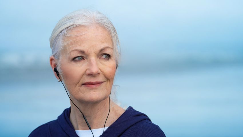 A woman over 50 with headphones in and the sea behind her